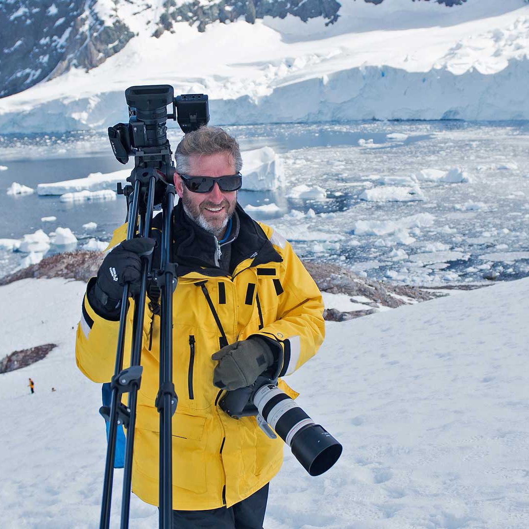 patrik_olofsson_antarktis Patrik Olofsson står i gul jacka bland snö och is på Antarktis med filmkamera