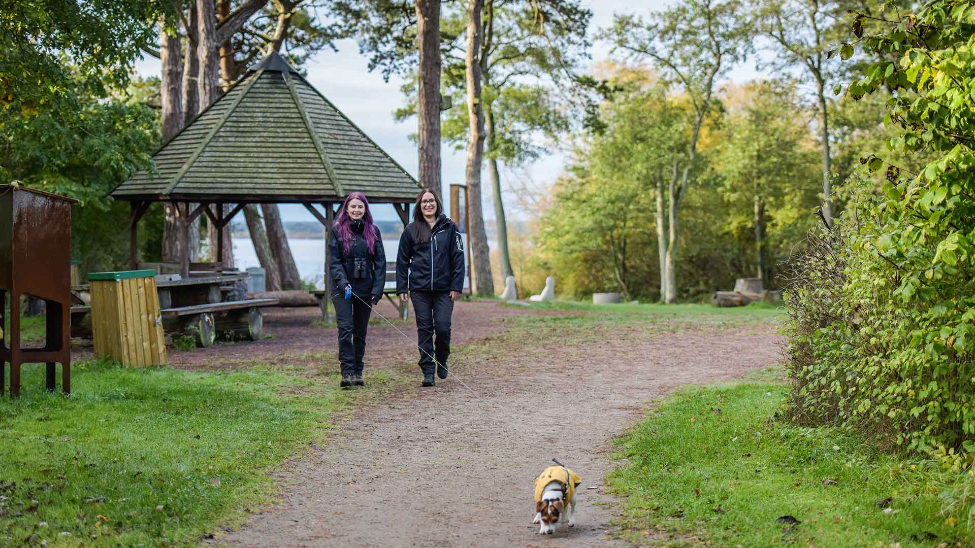 Två kvinnor och en liten hund promenerar på Ekenabben. I bakgrunden syns en paviljong och Hammarsjön. Foto Jennie Ahlqvist