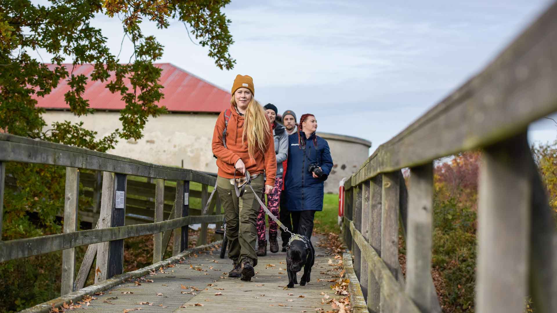 Fyra personer och en hund går över en träbro. I bakgrunden skymtar Lillö borgruin. Foto Jennie Ahlqvist.