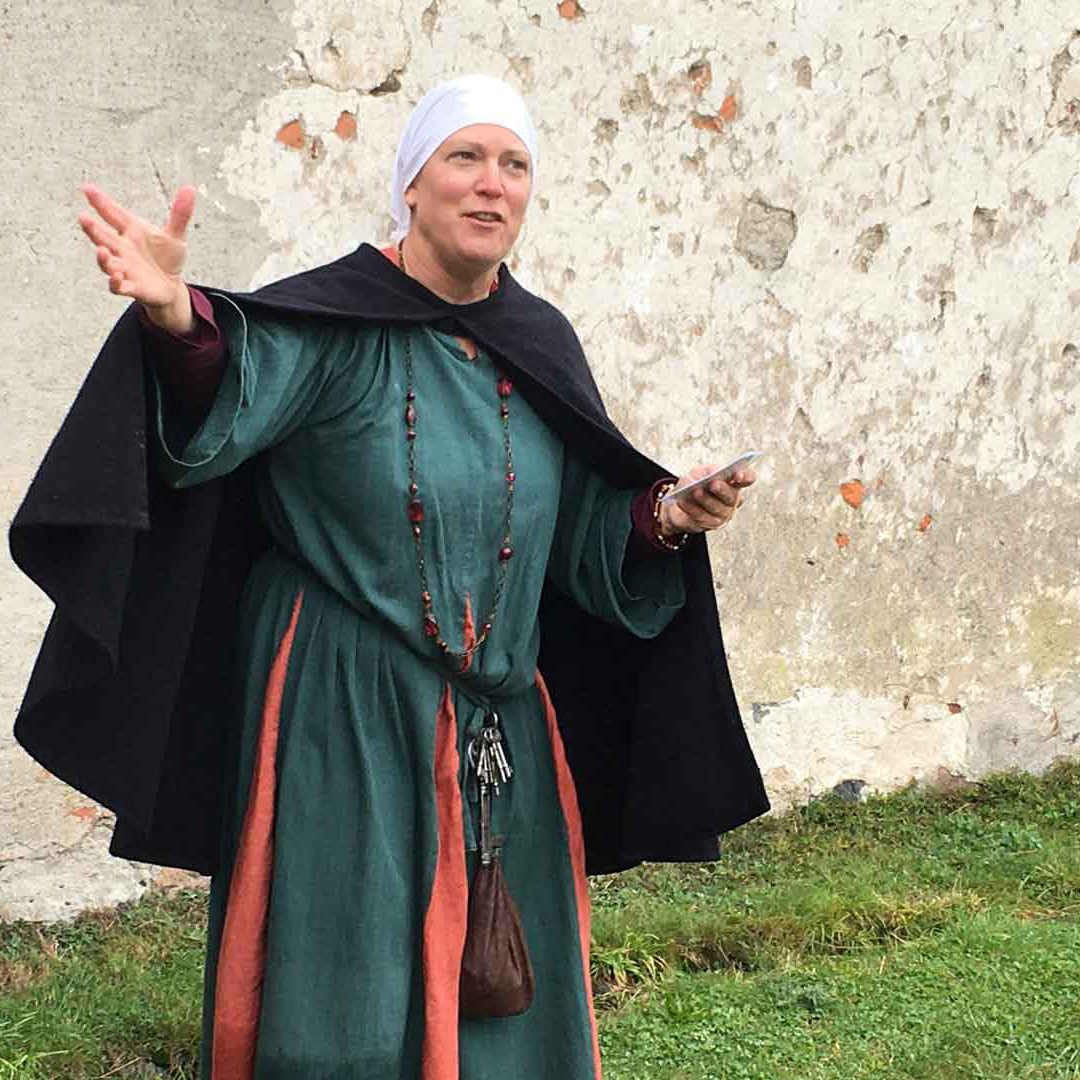 Kvinna i medeltidskläder står framför Lillö borgruin med utsträckta händer