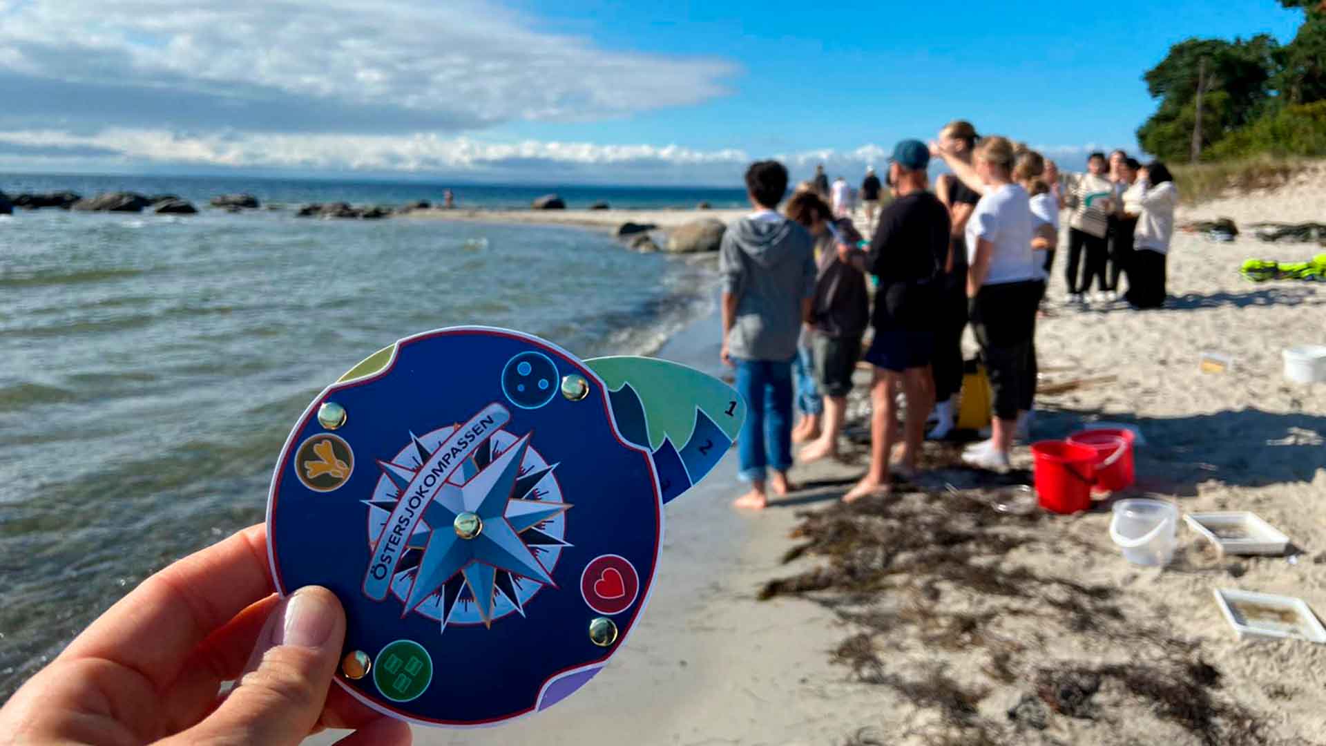 I förgrunden syns en färgglad papperssnurra med texten Östersjökompassen. I bakgrunden står en grupp ungdomar vid strandkanten.
