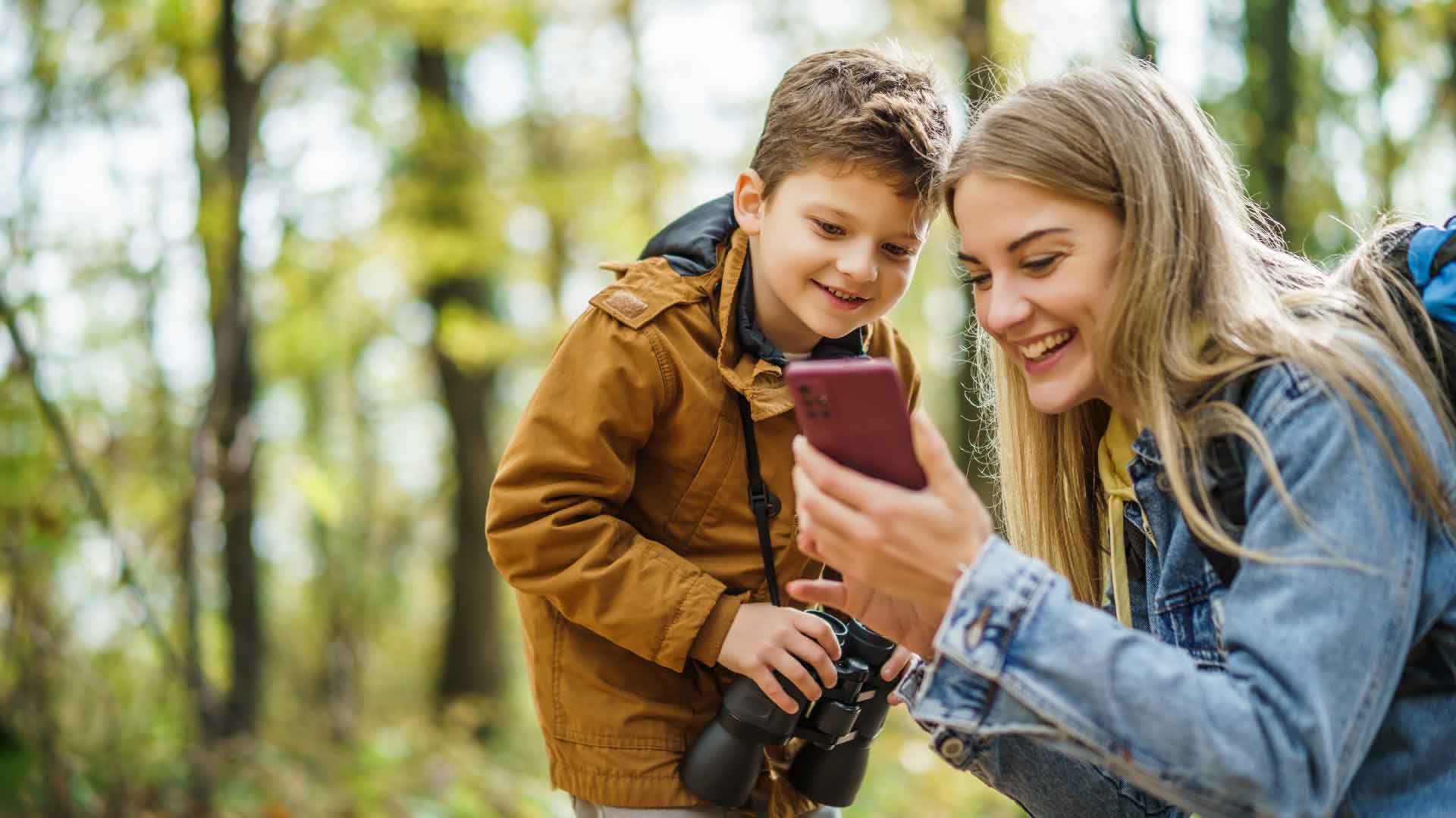 En glad mamma och ett barn med kikare tittar tillsammans på en mobil i skogen