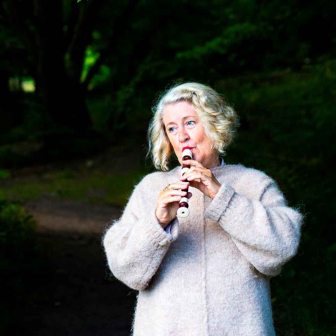 En kvinna står i grönskande lövskog och spelar flöjt.