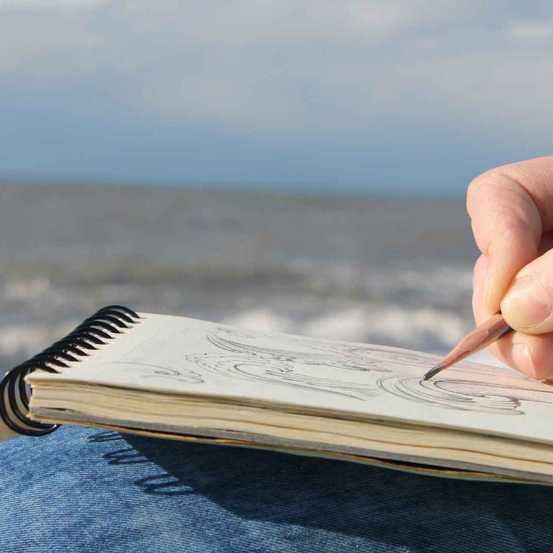 teckna_havet_AS Ett ritblock och en hand med en penna. I bakgrunden syns havet. Foto adoben Stock