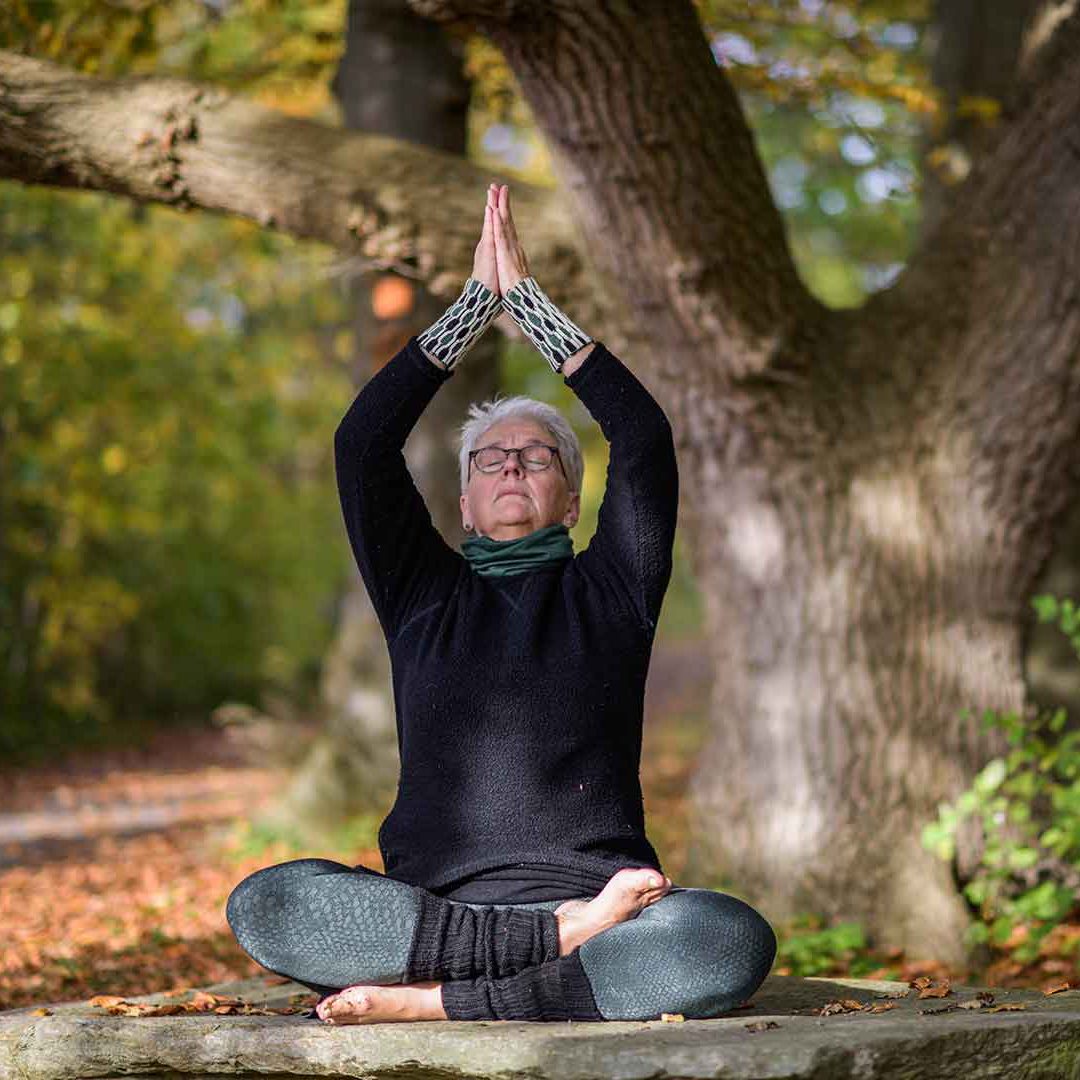 En kvinna sitter i skräddarställning med uppsträckta armar och gör yoga i lövskog. Foto Jennie Ahlqvist.