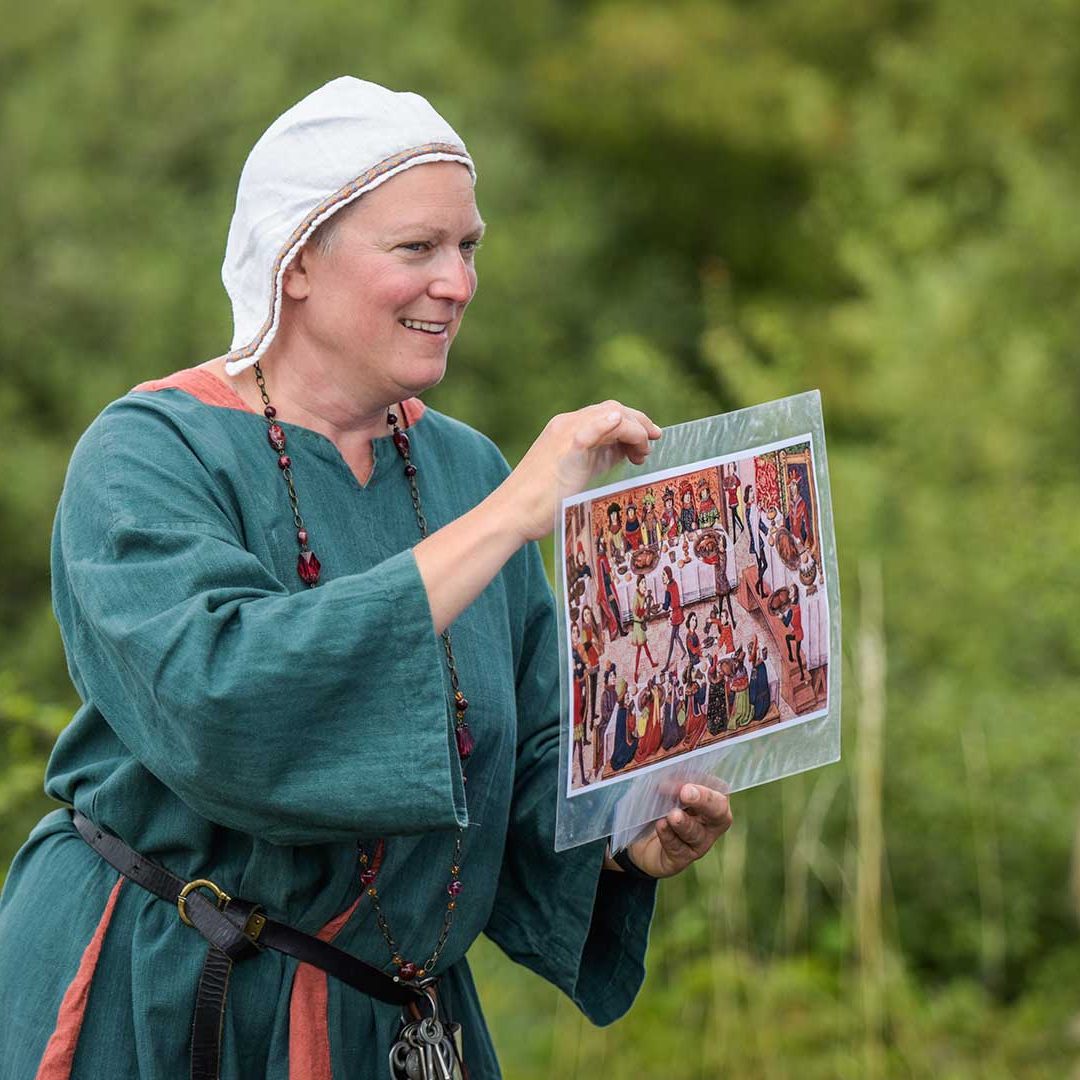 En medeltidsklädd kvinna håller upp en bild på ett medeltida gästabud.