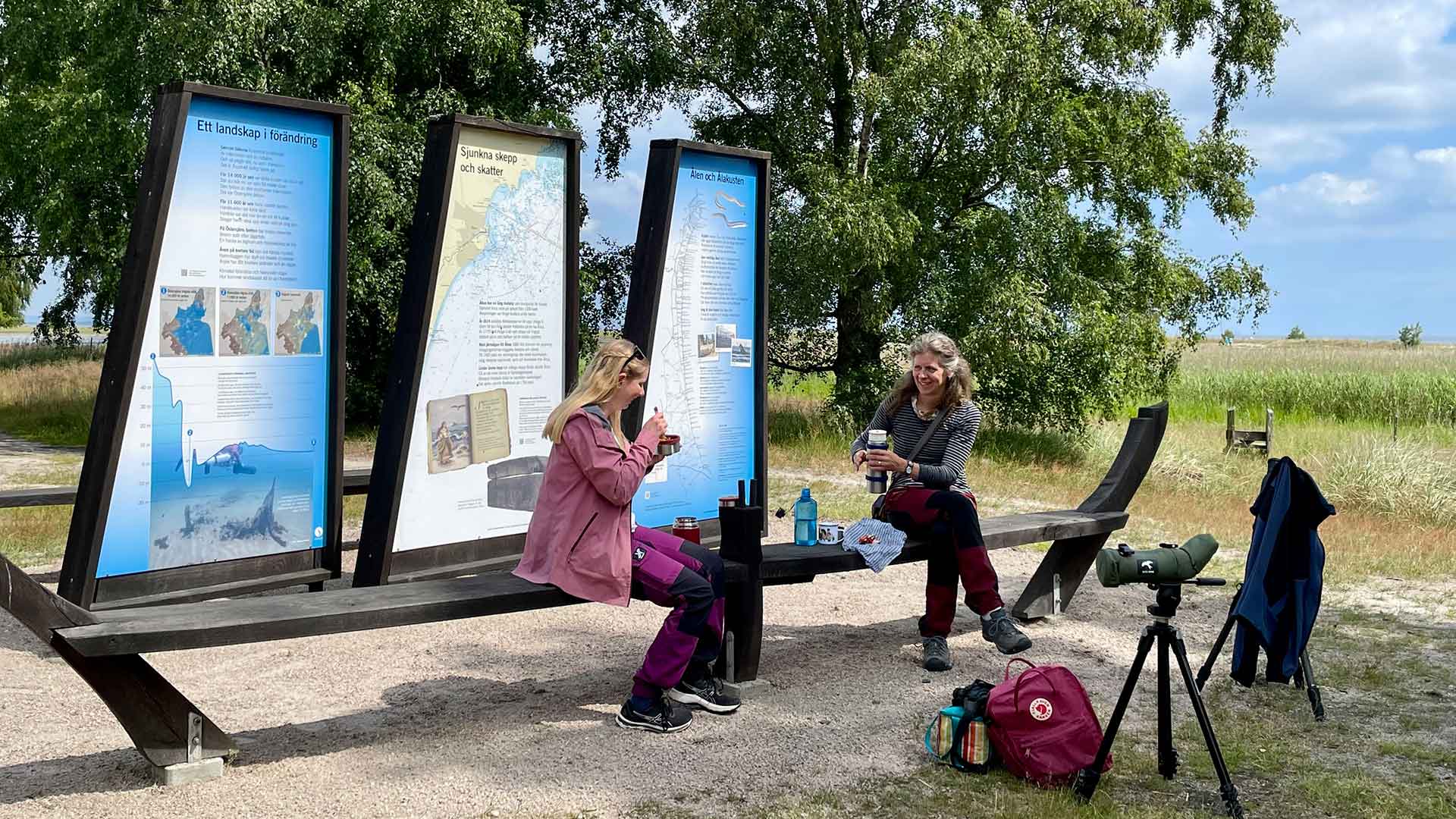 Två kvinnor sitter och fikar vid besöksplats Äspet, som består av tre segelskärmar och två bänkar i närheten av stranden. Foto Åsa Pearce