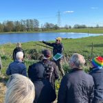 En grupp personer står och lyssnar till en man framför en våtmark.